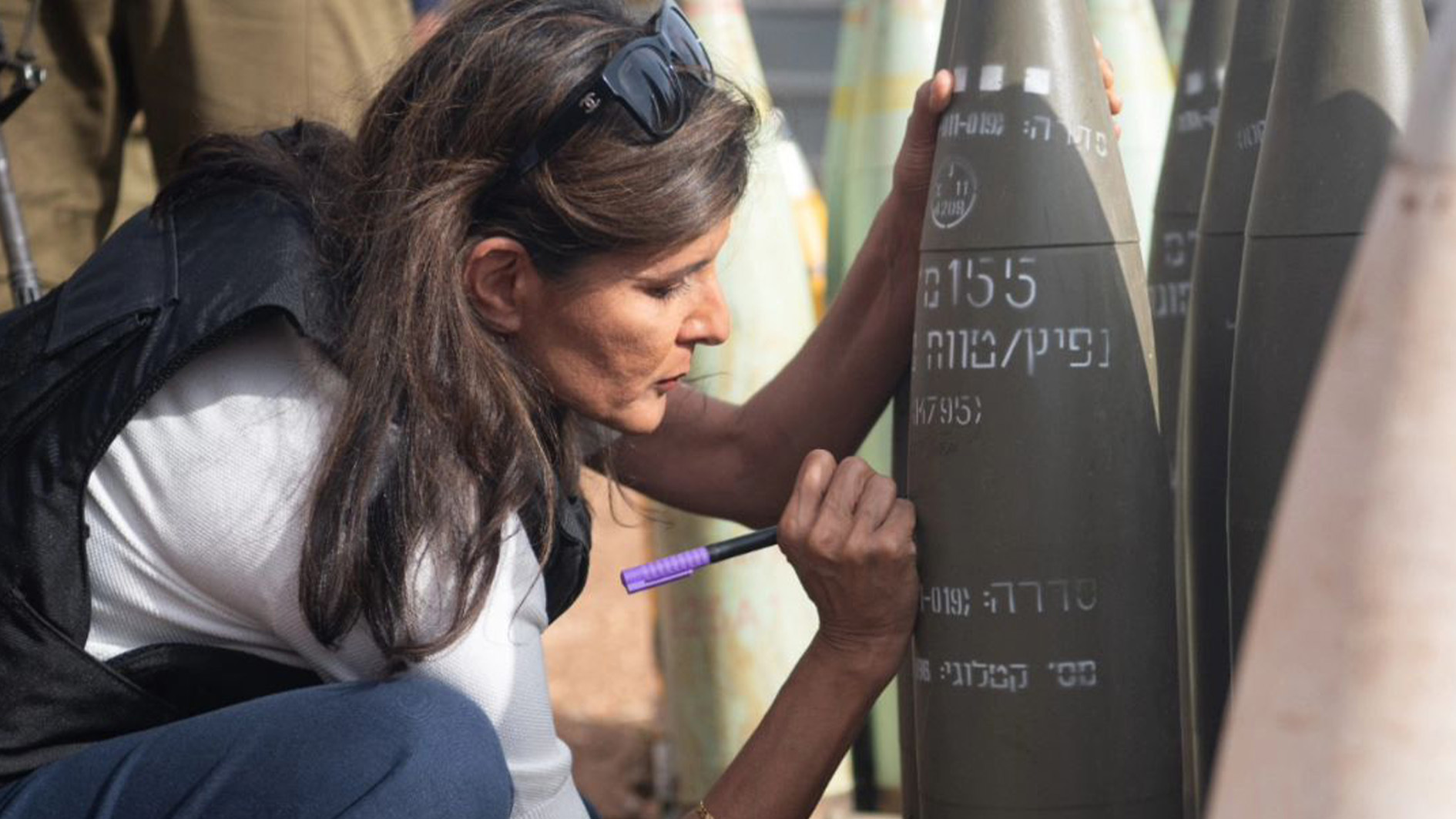 Side shot of Nikki Haley squatting down in a
vest signing an artillery shell